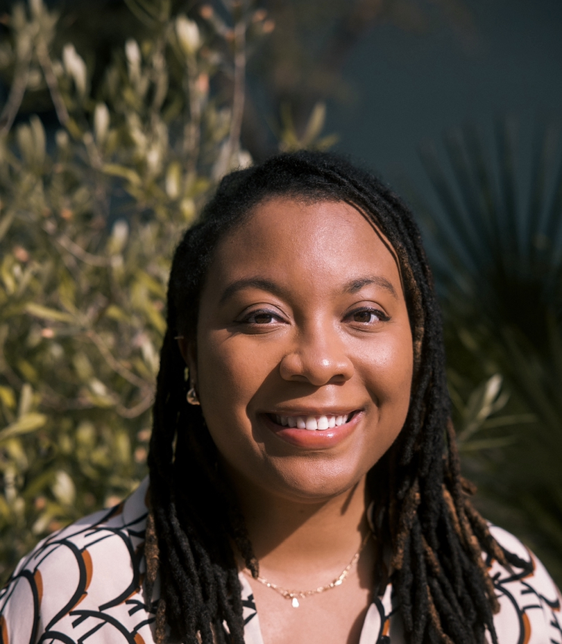 Dr. Rowe wears a printed button down shirt. She has locs and light brown skin. She smiles and stands in front of green bushes.